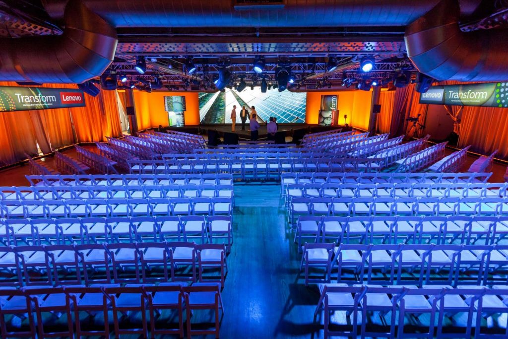 A brightly lit conference hall with empty chairs and a stage set for an event, featuring branding for 'transform' and 'lenovo.'.