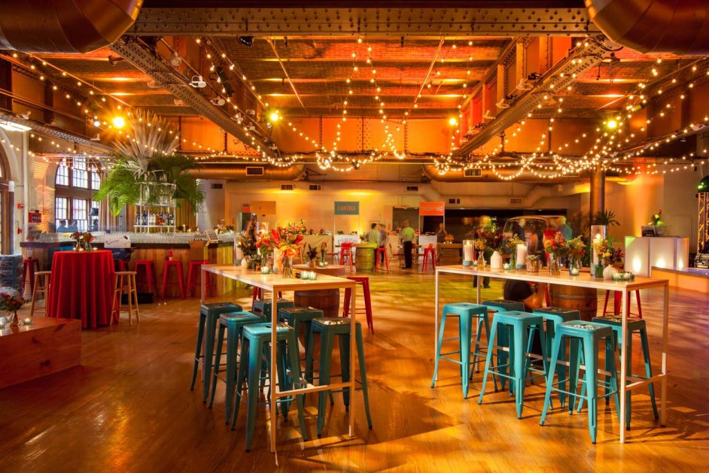 Spacious event hall adorned with string lights and floral centerpieces, featuring a bar and high stools.