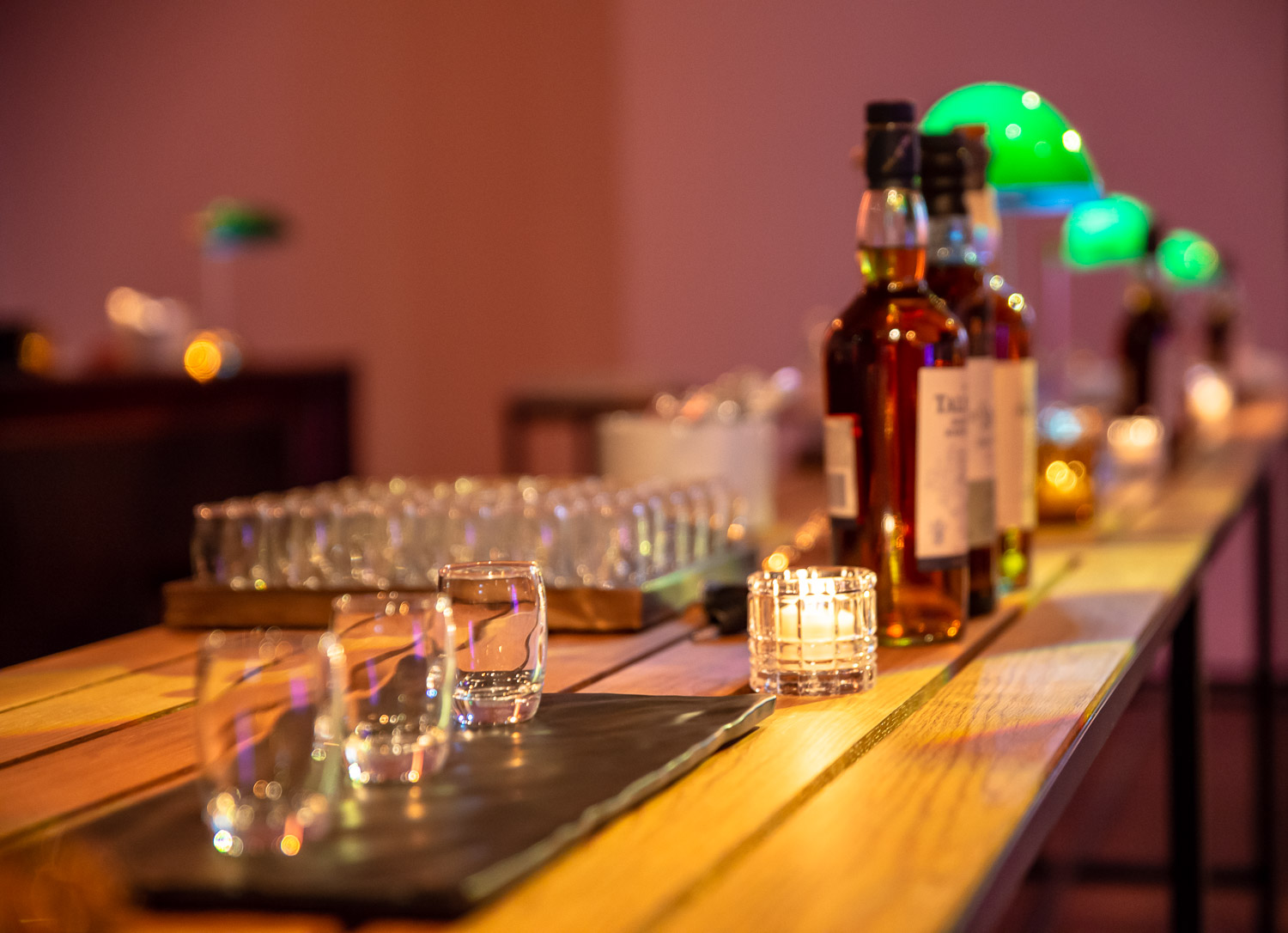 A whiskey tasting setup with bottles, glasses, and candles on a wooden table, with soft lighting in the background.