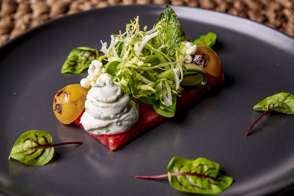 Gourmet dish featuring a slice of red meat topped with a dollop of herbed cream, garnished with greens, berries, and flowers, served on a dark plate.