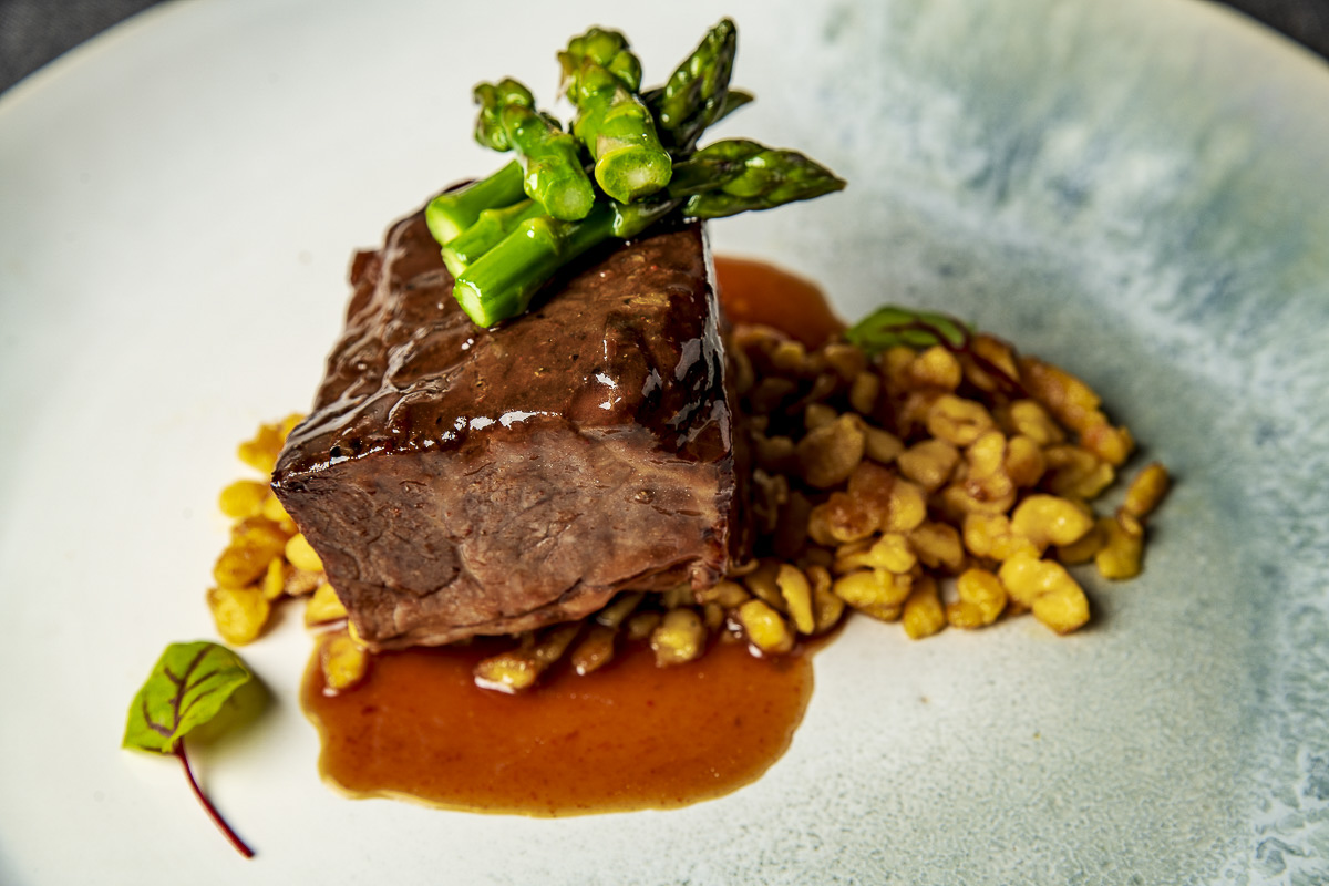 Filet mignon topped with asparagus, served with a side of corn and a rich sauce on a speckled plate.