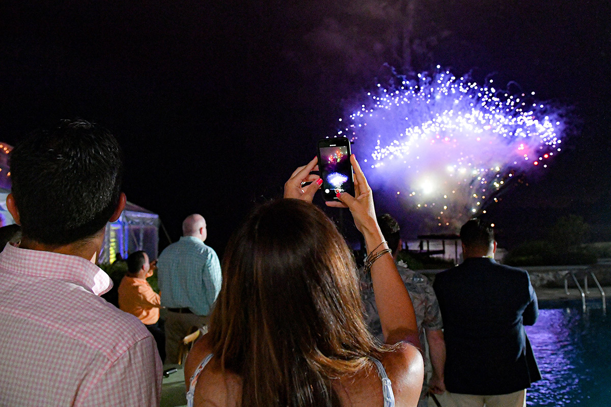 Person capturing fireworks on a smartphone camera at a gathering by the water.