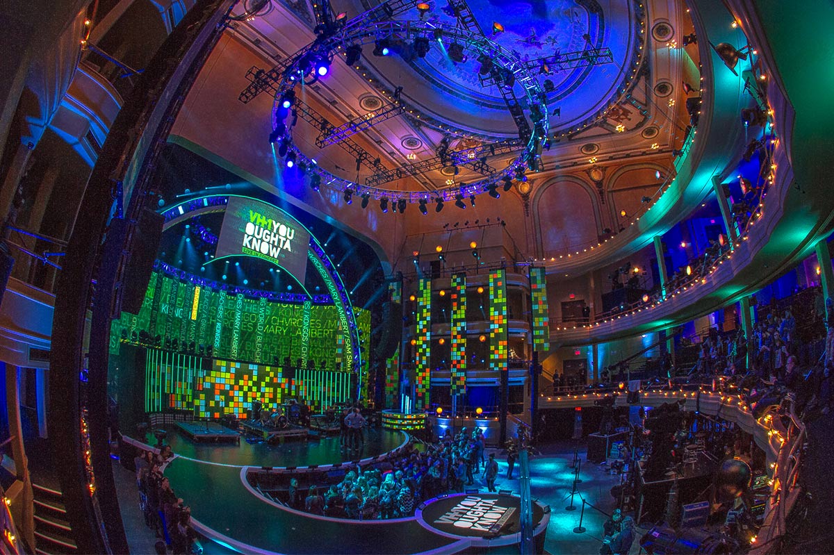 A wide-angle view of a vibrant and modern theater interior during a live event with an audience, featuring a large, illuminated stage with digital screens.